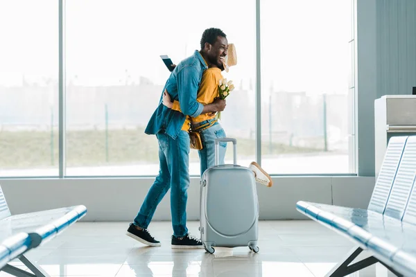 Щасливий афроамериканський чоловік з букетом тюльпанів, що обіймає дівчину з валізою та паспортом в аеропорту — стокове фото