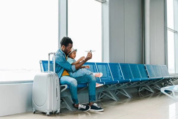 Afrikanisch-amerikanischer Vater und Sohn sitzen mit Koffer im Flughafen und spielen mit Spielzeugflugzeug — Stockfoto