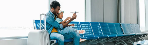 Plano panorámico de padre e hijo afroamericano sentado con maleta en el aeropuerto y jugando con avión de juguete - foto de stock
