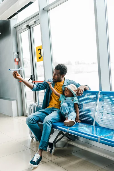Africano americano padre e hijo sentado en sala de espera en aeropuerto y jugando con juguete avión - foto de stock