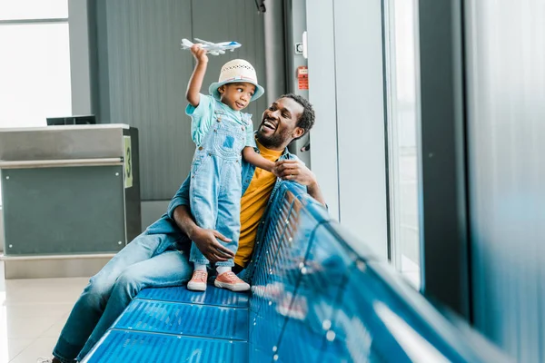 Heureux père afro-américain regardant son fils alors que le garçon joue avec un avion jouet à l'aéroport — Photo de stock