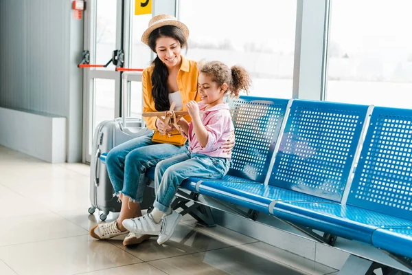 Feliz afroamericano madre e hija sentado en el aeropuerto con maleta y modelo de avión de madera - foto de stock