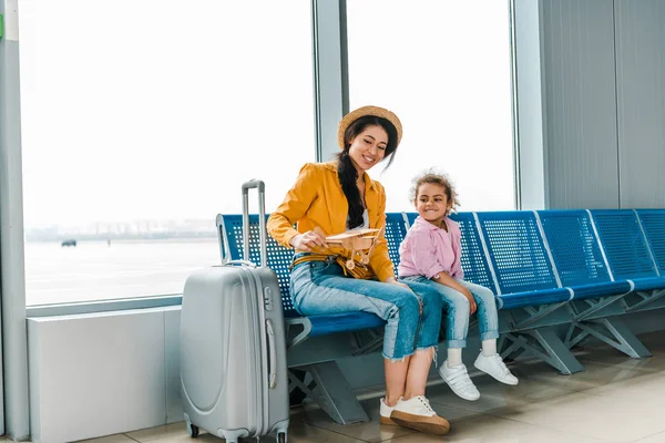 Sonriente afroamericano madre e hija sentado en el aeropuerto con maleta y modelo de avión de madera — Stock Photo