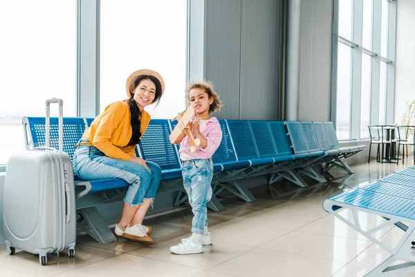 Happy african american mother and daughter in airport with suitcase and wooden plane model — Stock Photo