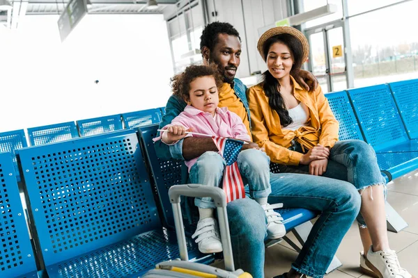 Familia afroamericana sentada en el aeropuerto con maleta mientras niño sosteniendo bandera americana - foto de stock