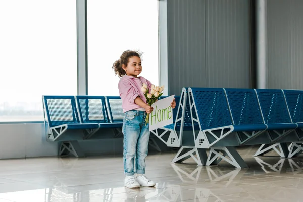 Веселий афроамериканець Kid стоячи в очікуванні залу в аеропорту і проведення тюльпани і плакат з домашнього написи — стокове фото
