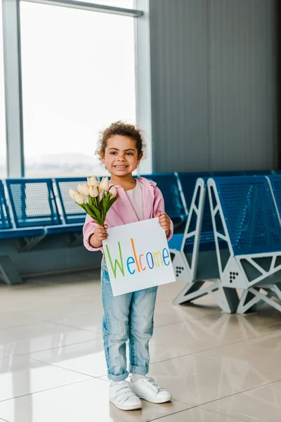 Happy african american kid standing in waiting hall in airport and holding tulips and placard with welcome lettering — Stock Photo