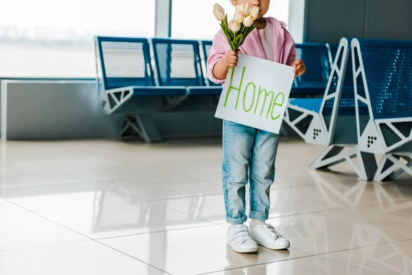 Vista recortada del niño afroamericano de pie en la sala de espera en el aeropuerto y la celebración de tulipanes y pancarta con letras en casa - foto de stock