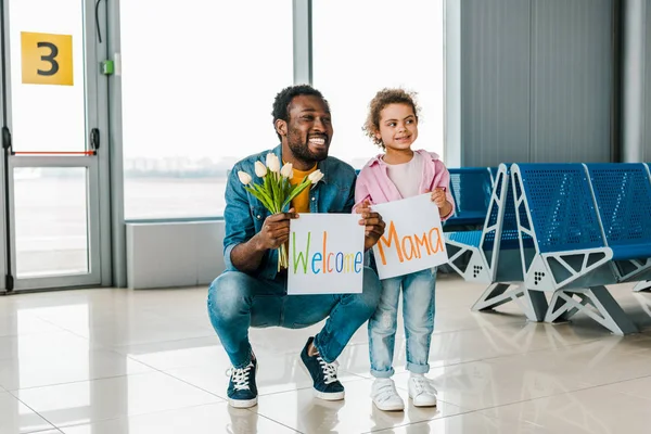 African american daughter and father standing in waiting hall in airport and holding tulips and placards with welcome mama words — Stock Photo
