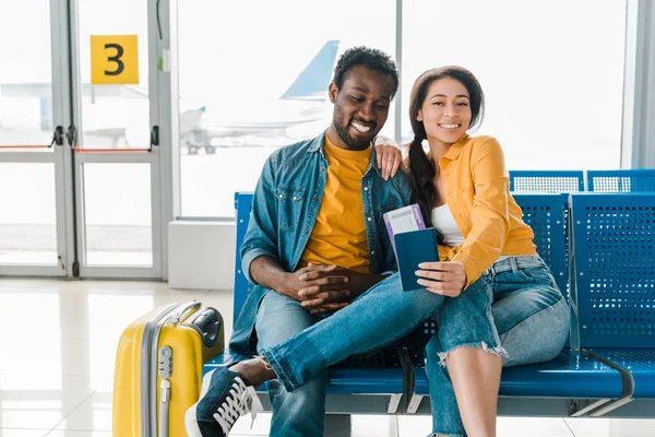 Feliz pareja afroamericana sentada en la sala de salida con maleta y billetes de avión en el aeropuerto - foto de stock