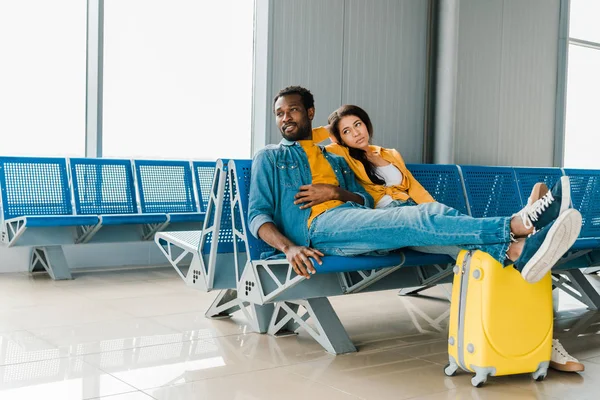 Pareja afroamericana cansada sentada en la sala de salida con maleta y esperando el vuelo - foto de stock