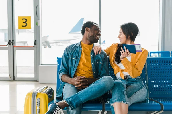 Feliz pareja afroamericana sentada en la sala de salida con maleta y billetes de avión y mirándose unos a otros - foto de stock