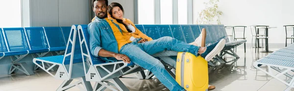 Plano panorámico de pareja afroamericana cansada sentada en la sala de salida con maleta y esperando el vuelo - foto de stock