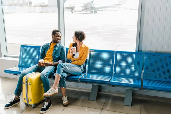 Feliz pareja afroamericana sentada en la sala de salida con café para llevar y maleta amarilla - foto de stock