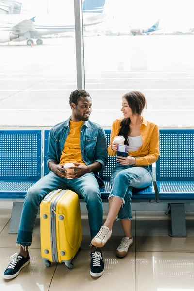 Улыбающаяся африканская американская пара, сидящая в зале вылета с кофе, чтобы пойти и желтый чемодан — стоковое фото