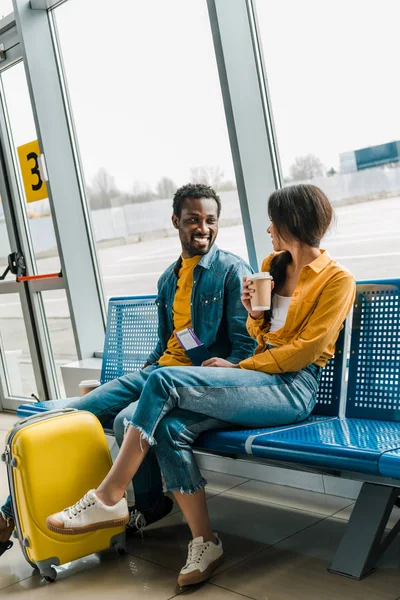 Feliz pareja afroamericana sentada en la sala de salida con café para llevar y maleta amarilla y mirándose - foto de stock
