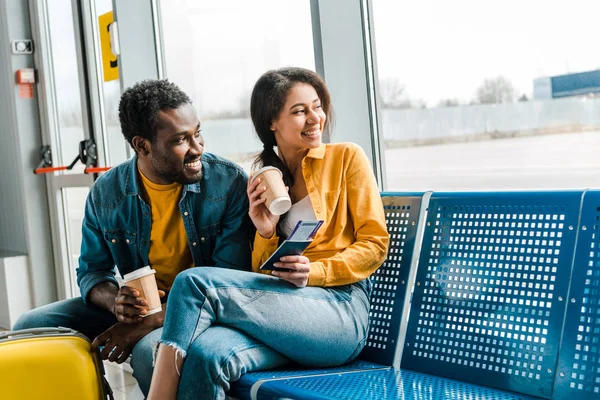 Heureux couple afro-américain assis dans le salon de départ avec café à emporter et billets d'avion et regarder la fenêtre — Photo de stock
