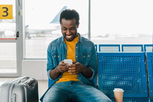 Улыбающийся африканский американец сидит в зале вылета и пользуется смартфоном — стоковое фото