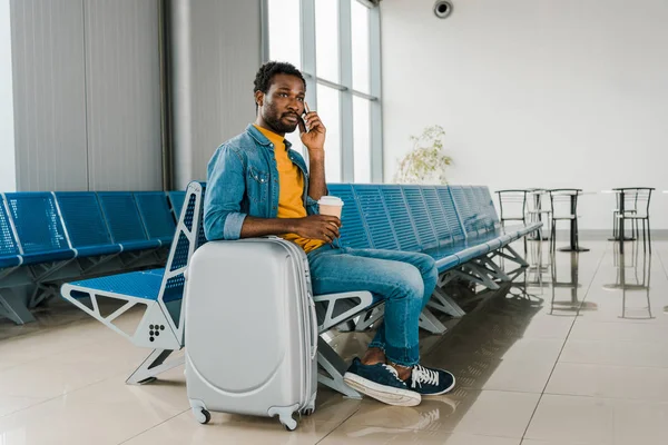 Homme afro-américain assis dans le salon de départ avec café pour aller et valise tout en parlant sur smartphone — Photo de stock