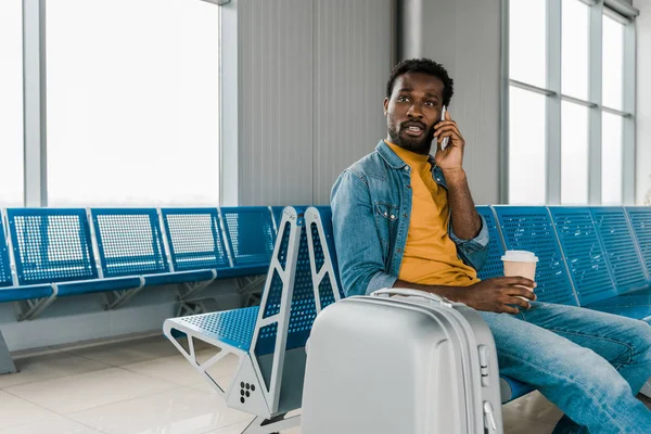 Homme afro-américain assis dans le salon de départ avec café pour aller et bagages tout en parlant sur smartphone — Photo de stock