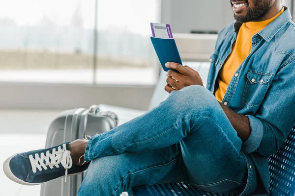 Vue recadrée d'un homme afro-américain souriant assis à l'aéroport avec billet d'avion et passeport — Photo de stock