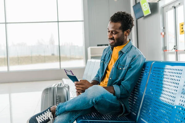 Hombre afroamericano sonriente sentado en el aeropuerto con billete de avión y pasaporte - foto de stock