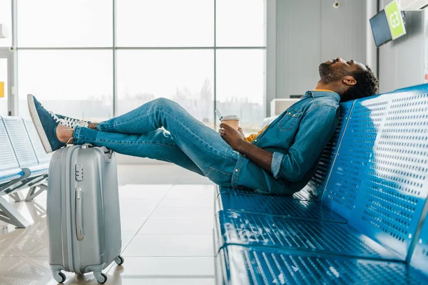 Africano americano hombre durmiendo en aeropuerto con café a ir y piernas en maleta - foto de stock