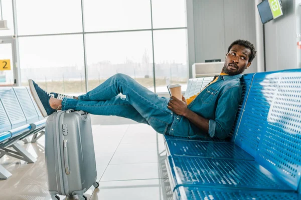 Hombre afroamericano sentado en el aeropuerto con café para llevar y piernas en la maleta - foto de stock