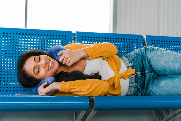 Улыбающаяся африканская американка лежит на сиденьях с подушкой для путешествий и закрытыми глазами в зале вылета — стоковое фото