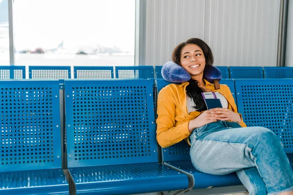 Sonriente mujer afroamericana sentada con almohada de viaje y pasaporte y billete de avión en la sala de salida - foto de stock