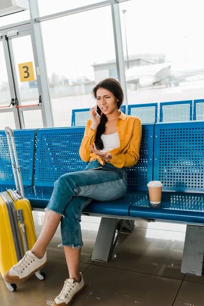 Mujer afroamericana insatisfecha sentada en salón de salida con maleta, café para llevar y hablar en smartphone - foto de stock