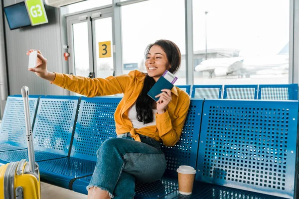 Femme afro-américaine souriante assise dans le salon de départ avec valise, café à emporter et selfie avec passeport et billet d'avion sur smartphone — Photo de stock
