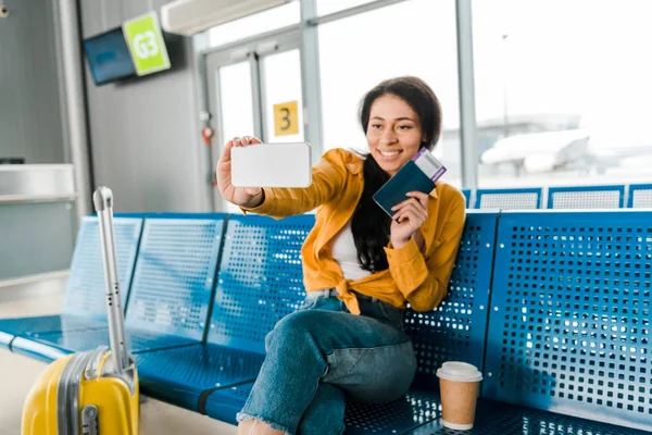 Счастливая африканская американка сидит в зале вылета с чемоданом, кофе, чтобы пойти и сделать селфи с паспортом и авиабилетом на смартфон — стоковое фото