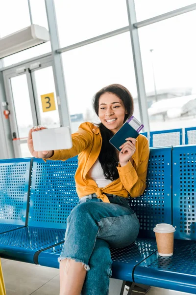 Heureuse femme afro-américaine assise dans le salon de départ et prenant selfie avec passeport et billet d'avion sur smartphone — Photo de stock