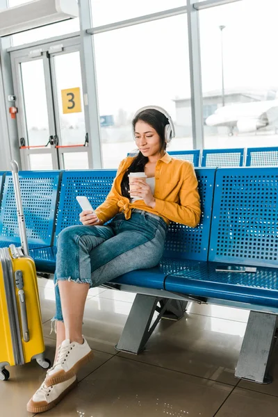 Happy african american woman sitting in departure lounge with coffee to go and suitcase while listening music in headphones and using smartphone — Stock Photo