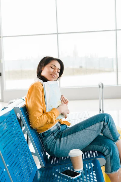 Sonriente mujer afroamericana sentada con café para ir y cerrar los ojos mientras sostiene el mapa en la sala de salida en el aeropuerto - foto de stock