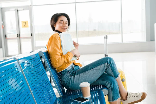 Zufriedene Afroamerikanerin sitzt mit Coffee to go und geschlossenen Augen in der Abflughalle des Flughafens und hält Karte in der Hand — Stockfoto