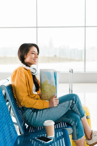 Sonriente mujer afroamericana sentada con café para llevar y teléfono inteligente en los asientos mientras sostiene el mapa en la sala de salida en el aeropuerto - foto de stock
