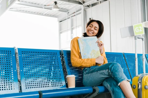 Улыбающаяся африканская американская женщина сидит с кофе, чтобы пойти и карта в зале вылета в аэропорту — стоковое фото