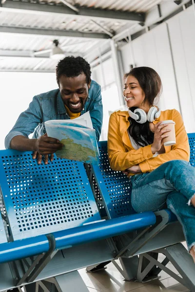 Sonriente mujer afroamericana con café para llevar y auriculares mirando novio feliz sosteniendo mapa en la sala de salida en el aeropuerto - foto de stock