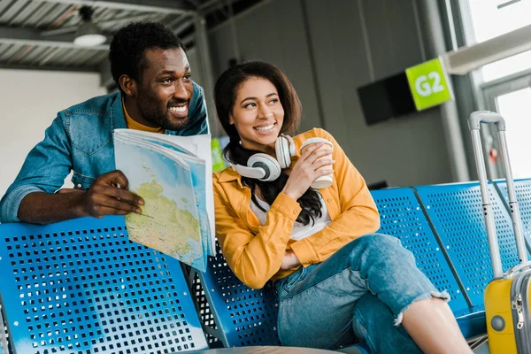 Sonriente pareja afroamericana sentada con mapa en la sala de salida en el aeropuerto y mirando hacia otro lado - foto de stock