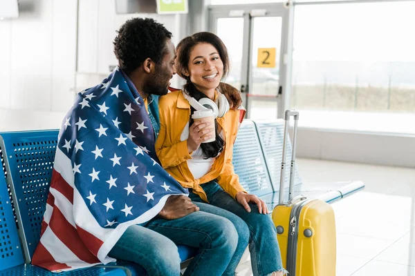 Feliz pareja afroamericana sentada con bandera americana en la sala de salida en el aeropuerto - foto de stock