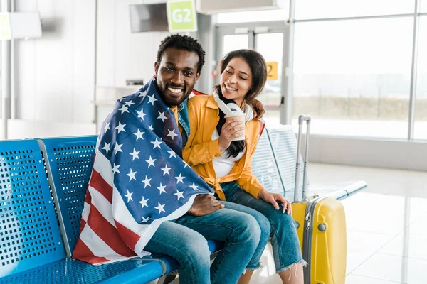 Felice coppia afroamericana seduta con bandiera americana e bagaglio nella sala partenze in aeroporto — Foto stock