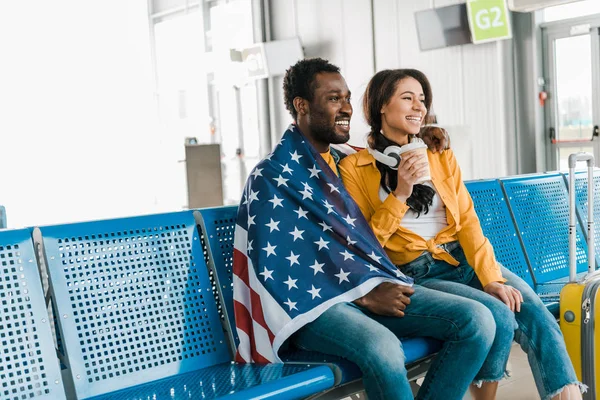 Feliz pareja afroamericana sentada con bandera americana y café para ir en la sala de salida en el aeropuerto - foto de stock