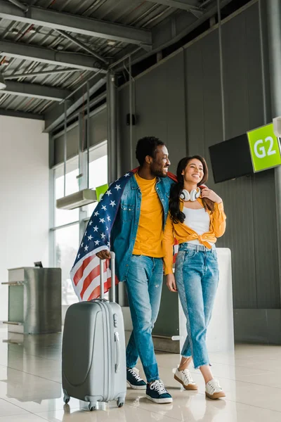 Feliz casal afro-americano com bandeira americana e mala andando na sala de embarque no aeroporto — Fotografia de Stock