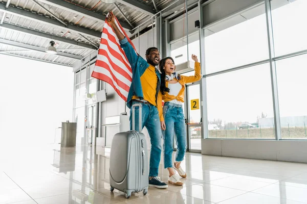 Glücklich aufgeregtes afrikanisch-amerikanisches Paar mit amerikanischer Flagge und Koffer in der Abflughalle des Flughafens — Stockfoto