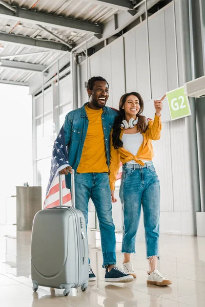 Sorrindo casal afro-americano com bandeira americana e mala andando na sala de embarque no aeroporto — Fotografia de Stock