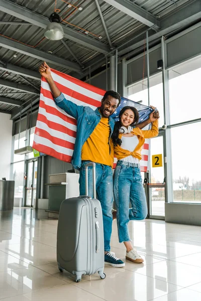 Sourire couple afro-américain avec drapeau américain et bagage marche dans le salon de départ à l'aéroport — Photo de stock