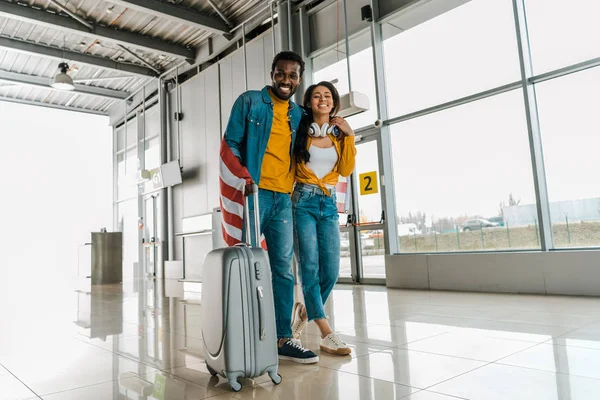 Glückliches afrikanisch-amerikanisches Paar mit amerikanischer Flagge und Gepäck zu Fuß in der Abflughalle des Flughafens — Stockfoto