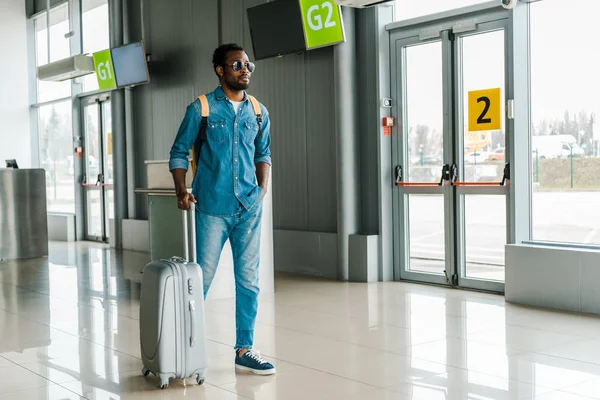 Handsome african american man standing with hand in pocket and suitcase in airport — Stock Photo